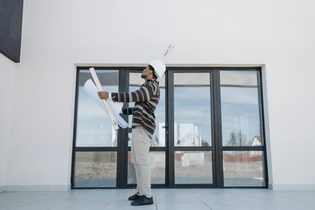 A black architect wearing a white helmet reviews construction plans outside a modern building.